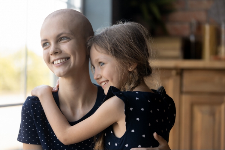 A young mother who is bald from chemotherapy is sitting in a kitchen holding her daughter. Both are smiling and looking out the window. Stem cell and bone marrow transplants can help people batting nearly 70 diseases, including blood cancer.