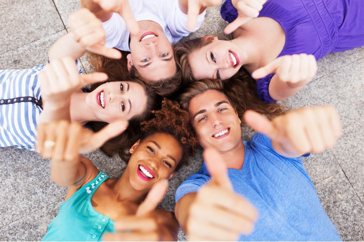 A group of diverse young people in their late teens/early 20s are lying in a circle and looking up at the camera, giving a thumbs-up.