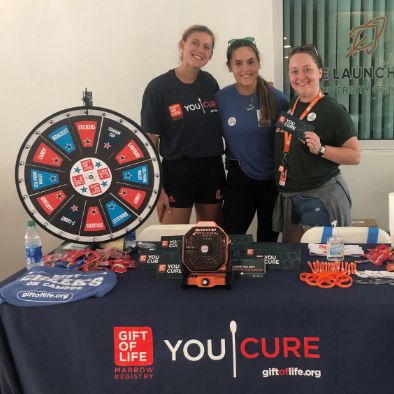 Three female college students who are Gift of Life Campus Ambassadors stand behind their swabbing table, which is laden with swab kits, orange wristbands, pens, and more.