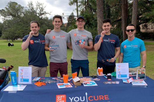 Five male college students wearing Gift of Life t-shirts stand behind a recruitment table set up with swab kits, pamphlets, and giveaways, as they invite fellow students to join the registry.