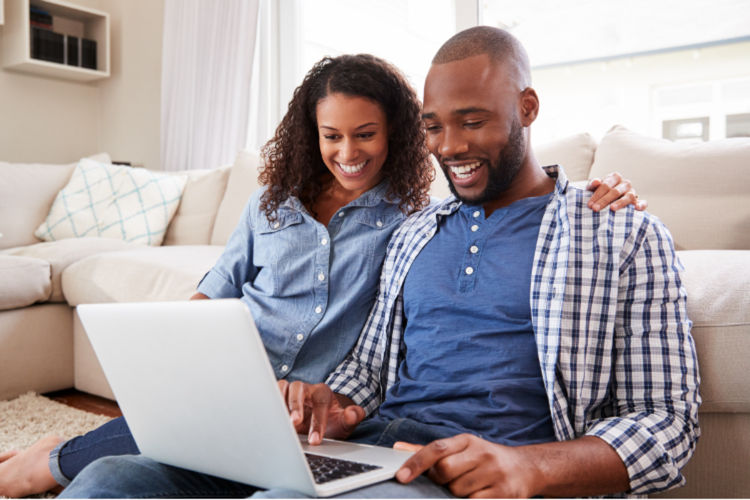 A Black couple in their thirties are sitting together on a couch and looking at a laptop computer. Both are smiling.