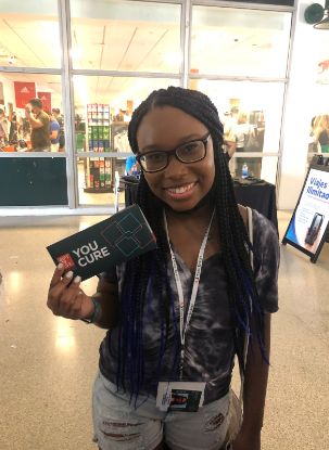 A smiling, Black, female college student is holding up a Gift of Life swab kit in front of her campus bookstore, as she encourages fellow students to join the marrow registry.