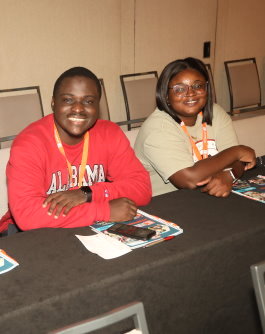 Two Gift of Life Campus Ambassadors sit at a table smiling during a break in the session. This Black male and female are smiling at the camera, and the young man is wearing a University of Alabama tee shirt. 
